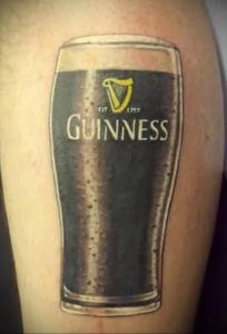 Фото тату на руке: реалистичное изображение бокала пива Guinness с логотипом арфы. Черно-серый рисунок татуировки.
