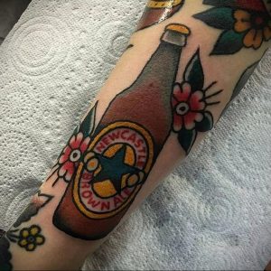 Фото тату на руке с изображением бутылки пива Newcastle Brown Ale, украшенной цветами. Яркий и красочный рисунок татуировки в стиле олдскул.