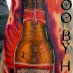 Фото тату на руке с изображением бутылки пива Budweiser, украшенной короной. Яркий и красочный рисунок татуировки в стиле реализм.