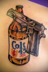 Фото тату на руке с изображением бутылки пива Colt 45 и пистолета. Реалистичный рисунок татуировки в черно-серых тонах.