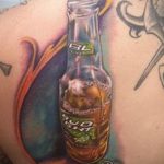 Фото тату на плече с изображением бутылки пива BL Lager. Реалистичный рисунок с использованием коричневых, желтых и оранжевых оттенков.
