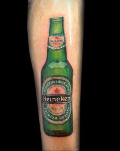 Фото тату на руке с изображением бутылки пива Heineken Lager. Детализированный рисунок в зеленых и золотых тонах.