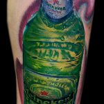 Фото тату на руке с реалистичным изображением бутылки пива Heineken. Детализированный рисунок с использованием зеленых, красных и белых оттенков.