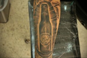 Фото тату на руке с изображением бутылки пива Corona в черно-белом стиле. Детализированный рисунок с надписью "Amigos".