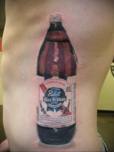 Фото тату на боку с реалистичным изображением бутылки пива Pabst Blue Ribbon. Детализированный рисунок с использованием синих, красных и белых оттенков.