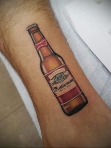 Фото тату с реалистичным изображением бутылки пива Budweiser на предплечье. Детализированный рисунок с использованием коричневых, красных и белых оттенков.