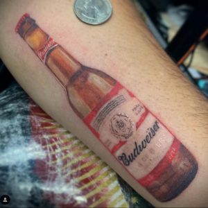 Фото тату: реалистичная татуировка бутылки пива Budweiser на руке. Детализированный рисунок татуировки с изображением этикетки и бликов света.