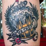 Фото тату на руке: бокал пива с надписью 'In Beer We Trust', окруженный колосьями пшеницы, выполненное в традиционном стиле.