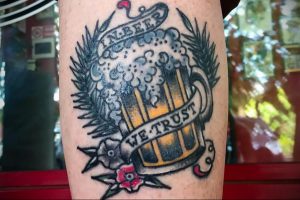 Фото тату на руке: бокал пива с надписью 'In Beer We Trust', окруженный колосьями пшеницы, выполненное в традиционном стиле.