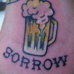 Фото тату кружки пива с надписью 'SORROW' под ней. Рисунок татуировки в стиле олдскул с использованием синих и желтых цветов.