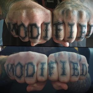 Фото татуировки на костяшках пальцев с надписью 'MODIFIED'. Черный цвет, простой шрифт, минималистичный дизайн.
