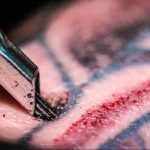 Крупный план процесса татуировки, где игла для татуировки касается кожи. Видны следы крови и синие линии уже нанесенной татуировки.