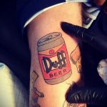 Фото тату Duff Beer в процессе нанесения на ногу. Видны руки мастера с тату-машинкой и контуры рисунка.