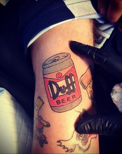 Фото тату Duff Beer в процессе нанесения на ногу. Видны руки мастера с тату-машинкой и контуры рисунка.