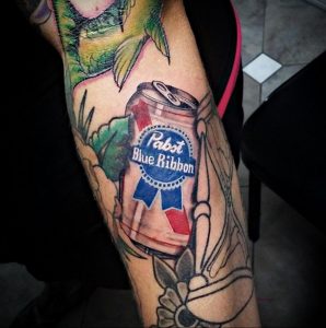 Фото тату банки пива Pabst Blue Ribbon на предплечье, выполненное в ярких цветах с элементами традиционного стиля.