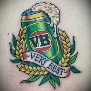 Фото тату на руке с изображением банки пива VB Very Best, окруженной колосьями пшеницы и лентой с надписью. Традиционный стиль.