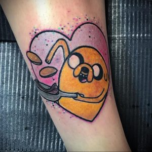 Фото тату персонажа Джейка из мультфильма Adventure Time и пончика в форме сердца на икре. Рисунок выполнен в ярких цветах с использованием контуров.