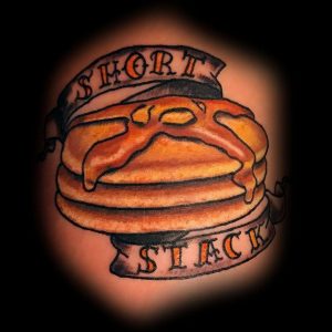 Фото татуировки с изображением стопки блинчиков и надписью «Short Stack» на фоне. Рисунок выполнен в традиционном стиле.