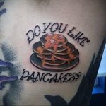 Фото татуировки с изображением стопки блинчиков и надписью «Do you like pancakes?» на плече. Рисунок выполнен в ярком, мультяшном стиле.