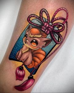 Фото тату Vulpix на предплечье. Оранжевый покемон Vulpix в рамке с кисточкой. Красочный рисунок татуировки в стиле аниме и японской гравюры.