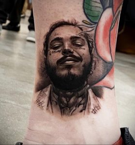 Фото тату реалистичного портрета Post Malone на предплечье, выполненного в черно-серых тонах с элементами цветных брызг.