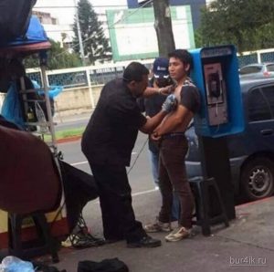 Фото тату процесса нанесения татуировки на улице. Мастер делает татуировку мужчине рядом с телефонной будкой. Небезопасные условия и отсутствие стерильности.