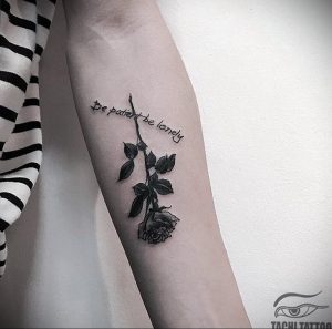 Фото тату: черная татуировка розы с надписью "Be patient be lonely" на предплечье. Элегантный рисунок с каллиграфическим шрифтом.