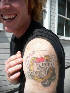 Фото тату: татуировка с изображением игральных карт, подковы и надписи 'Lucky Devil' на плече.