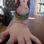 Фото тату: татуировка подковы с числом 25, выполненная в традиционном стиле, на запястье руки. Вид с другого ракурса.