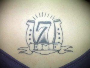 Фото тату цифры 7 с именем EMF на ленте. Простой рисунок татуировки в стиле олдскул с использованием черного цвета.
