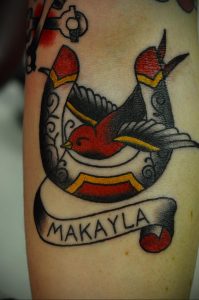 Фото тату ласточки с именем 'Makayla' на ленте. Классический рисунок татуировки в стиле олдскул с использованием красного, черного и желтого цветов.