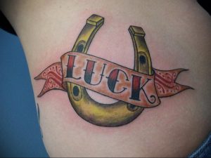 Фото тату на боку: изображение двух золотых подков и надписи "LUCK" на ленте, выполненное в ярких цветах в стиле олд скул.