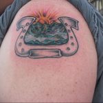 Фото татуировки подковы, внутри которой изображен горный пейзаж с восходом солнца. Рисунок выполнен в реалистичном стиле. Расположение на плече.