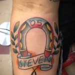Фото татуировки подковы с именем 'Steven' и датой '05' на ленте, выполненной в ярком и красочном стиле.
