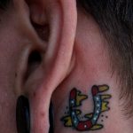 Фото миниатюрной татуировки подковы за ухом, выполненной в ярком и красочном стиле с использованием желтого, красного и синего цветов.