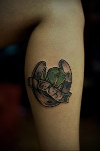 Фото тату подковы с инициалами 'G.M.A.M' на ленте, выполненное в реалистичном стиле с использованием мягких оттенков.