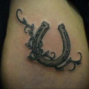 Фото тату подковы, украшенной растительным орнаментом в графичном стиле. Черно-белый рисунок татуировки на теле.