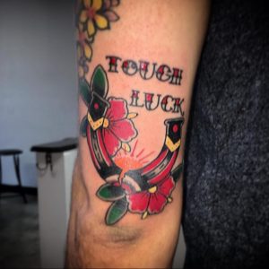 Фото тату подковы с красными цветами и надписью 'TOUGH LUCK' на руке. Цветная татуировка в стиле олдскул с яркими деталями.