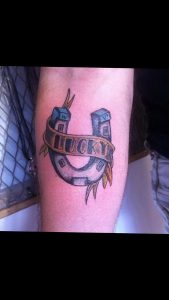 Фото тату подковы с надписью 'Lucky' на предплечье. Черно-серый рисунок татуировки в стиле олдскул с яркими акцентами.
