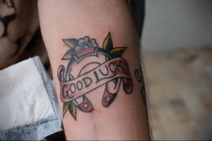 Фото татуировки подковы с надписью "Good Luck" в стиле олд скул на предплечье. Яркие цвета, четкие контуры, традиционные элементы: цветы, лента. Классический морской стиль.