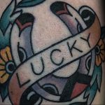 Фото тату на руке: подкова с надписью 'LUCKY', украшенная цветами и лентами. Яркие цвета, стиль олдскул.
