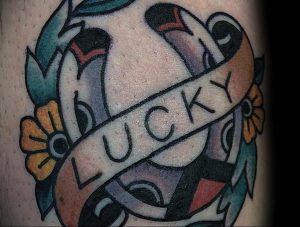 Фото тату на руке: подкова с надписью 'LUCKY', украшенная цветами и лентами. Яркие цвета, стиль олдскул.