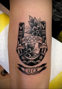 Фото тату подковы с цветами и надписью "GOOD LUCK" в черно-белом стиле. Четкие линии и детализированная прорисовка.