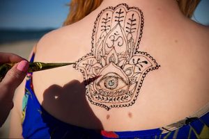 Фото хенна-татуировки Хамса (Рука Фатимы) на спине, украшенной сложным узором и изображением глаза. Выполнена в коричневых тонах.