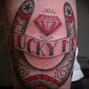Фото тату подковы с крупным бриллиантом и надписью 'LUCKY ME' в стиле олд скул на руке. Яркий рисунок татуировки с использованием красного, черного и серого цветов.