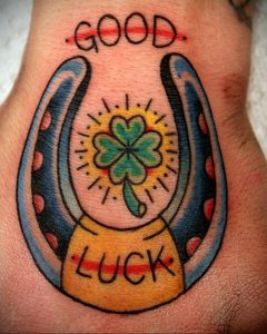 Фото тату на запястье: подкова, клевер и надпись 'Good Luck' в стиле олд скул с яркими цветами и четкими линиями.