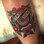 Фото тату на ноге: подкова, окруженная цветами и лентой с надписью "Stay Lucky". Яркий и символичный рисунок в стиле олдскул.