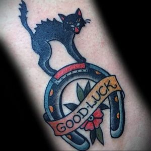 Фото тату на руке: черная кошка сидит на подкове с надписью "GOOD LUCK". Яркий и символичный рисунок в стиле олдскул.