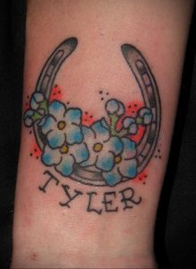 Фото тату подковы с голубыми цветами и надписью 'Tyler'. Яркий рисунок, персонализированный дизайн, символ удачи и памяти.