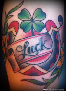 Фото тату на предплечье: яркий клевер с четырьмя листьями над надписью 'Luck' в стиле олд скул. Красные, зеленые и черные цвета.